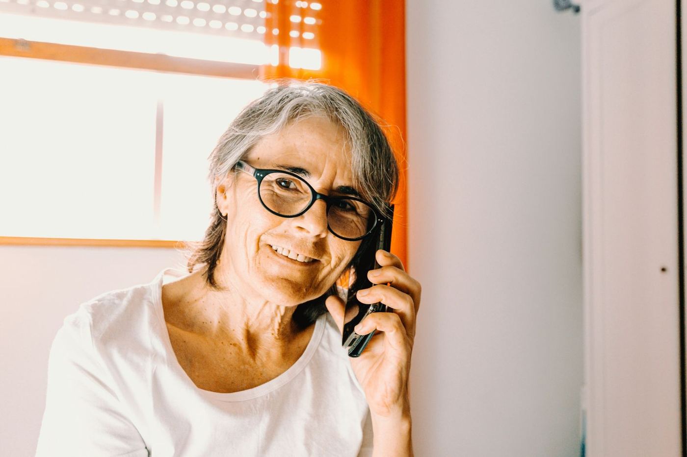 An older woman in glasses, smiling as she talks on her mobile phone at home.
