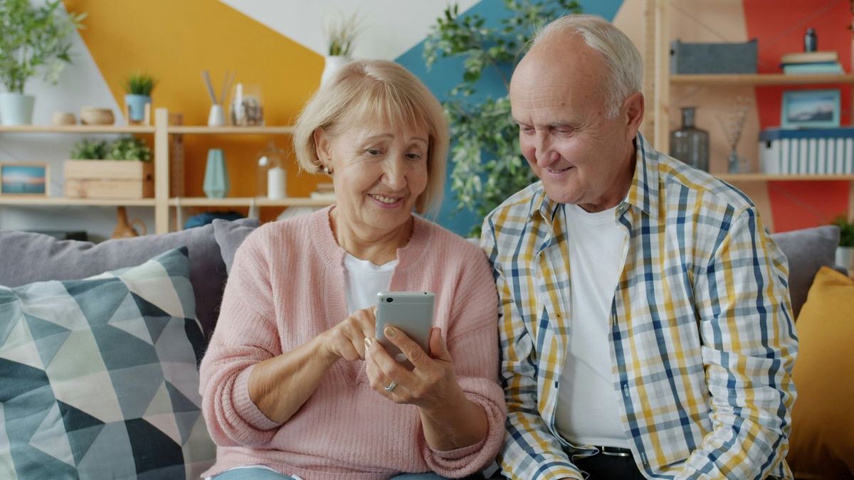An older couple on a sofa, smiling as they look at a smartphone together.